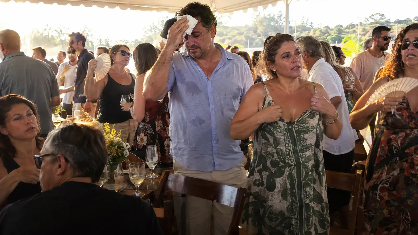 Convidados de uma festa sob tenda sofrendo com o calor: um homem enxuga o suor da testa com um guardanapo e mulheres se abanam com leques