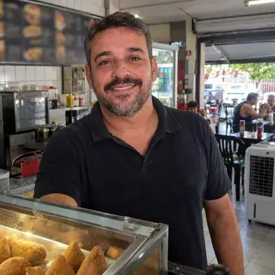 Eduardo, dono de lanchonete, de camisa polo preta atrás do balcão de salgados, com clientes nas mesas e um climatizador branco ao fundo