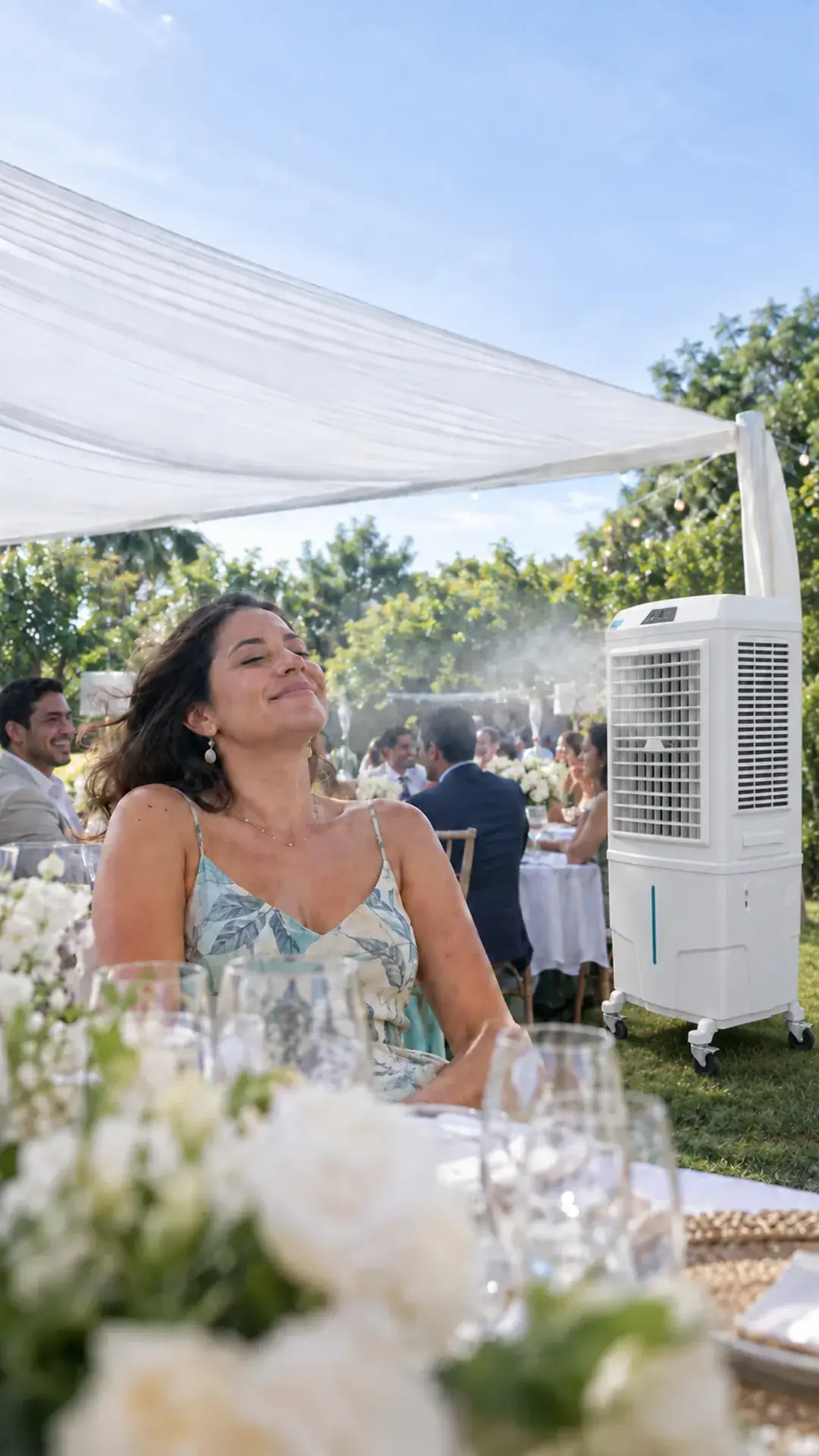 Convidada de vestido estampado de folhas sorri de olhos fechados à mesa de uma festa ao ar livre, sob tenda branca e entre arranjos de flores brancas, enquanto um climatizador branco com rodízios sopra névoa fresca sobre os convidados no gramado.
