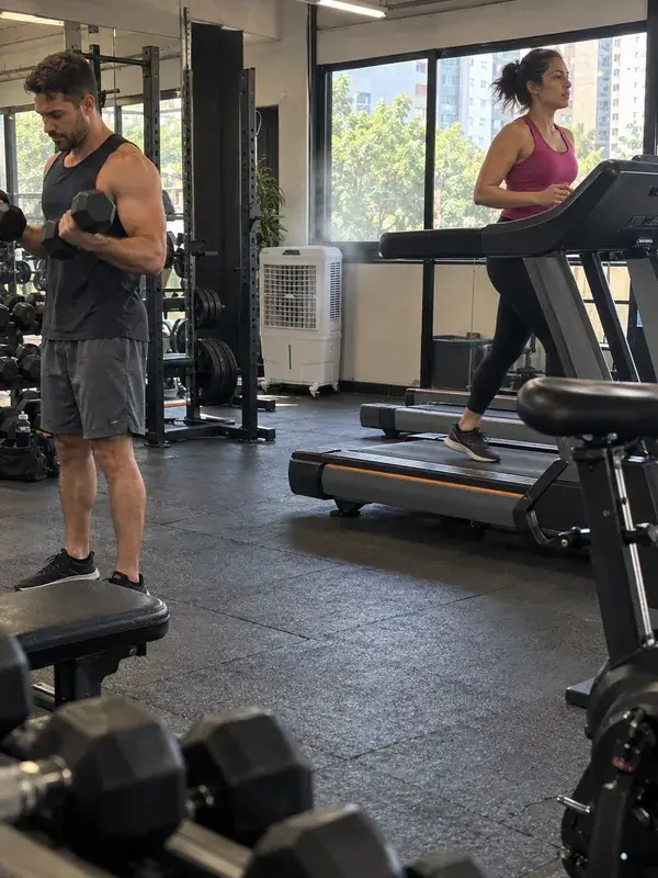 Homem treinando com halteres e mulher correndo na esteira em uma academia, com um climatizador branco ligado perto das janelas