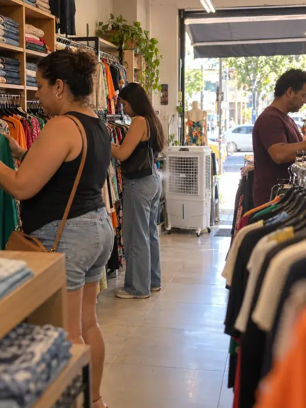 Clientes escolhendo roupas nas araras de uma loja com a porta aberta para a rua, com um climatizador branco ligado perto da entrada
