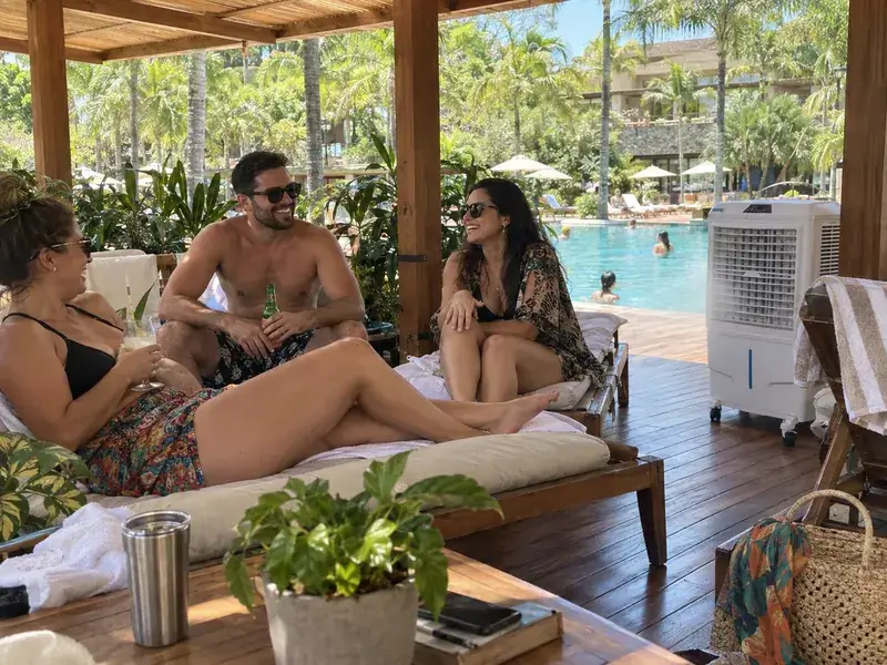 Hóspedes conversando em espreguiçadeiras de um deck coberto à beira da piscina de um hotel, com um climatizador branco ao lado