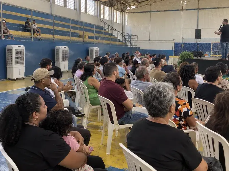 Público sentado em cadeiras plásticas assistindo a uma palestra em um ginásio municipal, com três climatizadores brancos junto à parede lateral