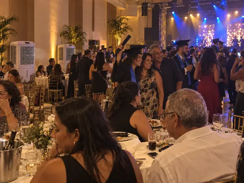 Formanda de beca erguendo o canudo ao lado dos pais em uma festa de formatura com palco iluminado, com dois climatizadores brancos junto à parede