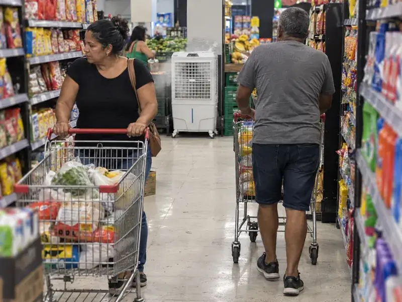 Clientes empurrando carrinhos por um corredor de supermercado, com um climatizador branco ligado no fim do corredor, perto do hortifrúti